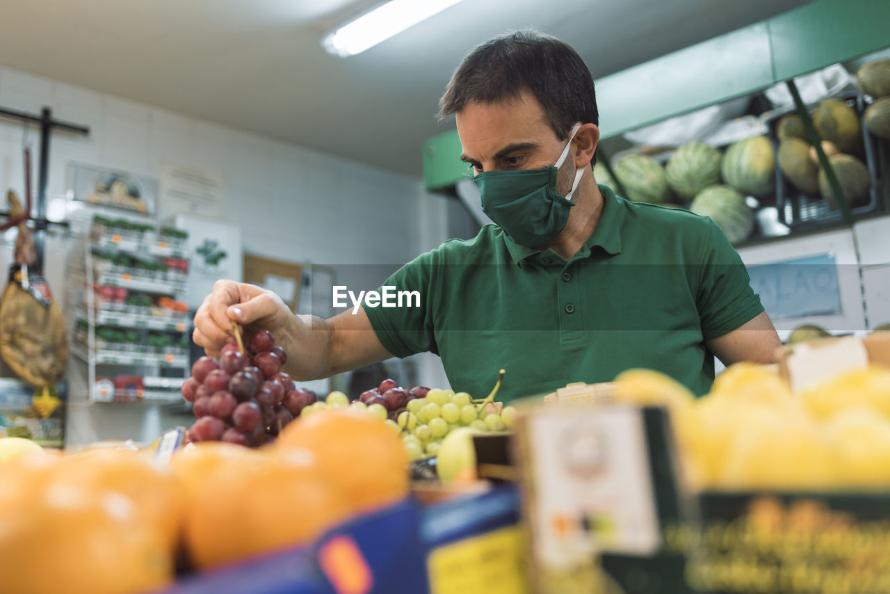 A man wearing a mask picking up a bunch of black grapes