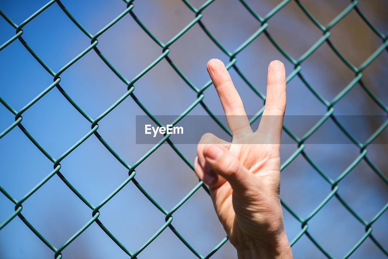 Cropped hand gesturing peace sign by green chainlink fence
