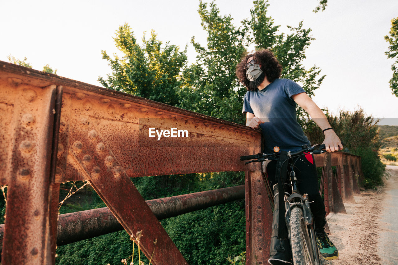 Man wearing mask standing with bicycle on wooden bridge
