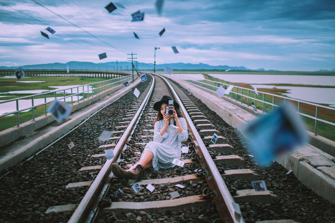 Full length of young woman photographing from camera while sitting amidst instant print transfers on railroad track