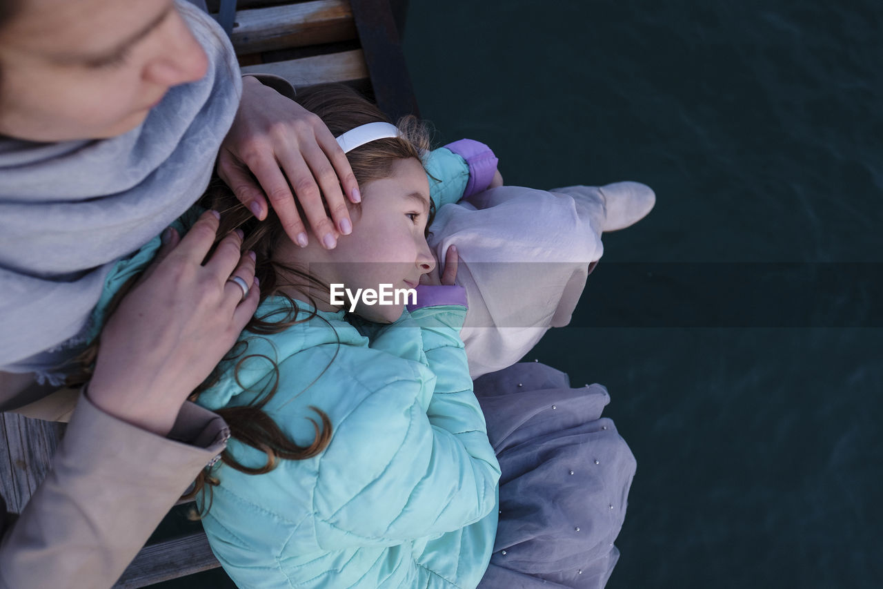 High angle view of daughter lying on mother's lap while sitting on pier over sea