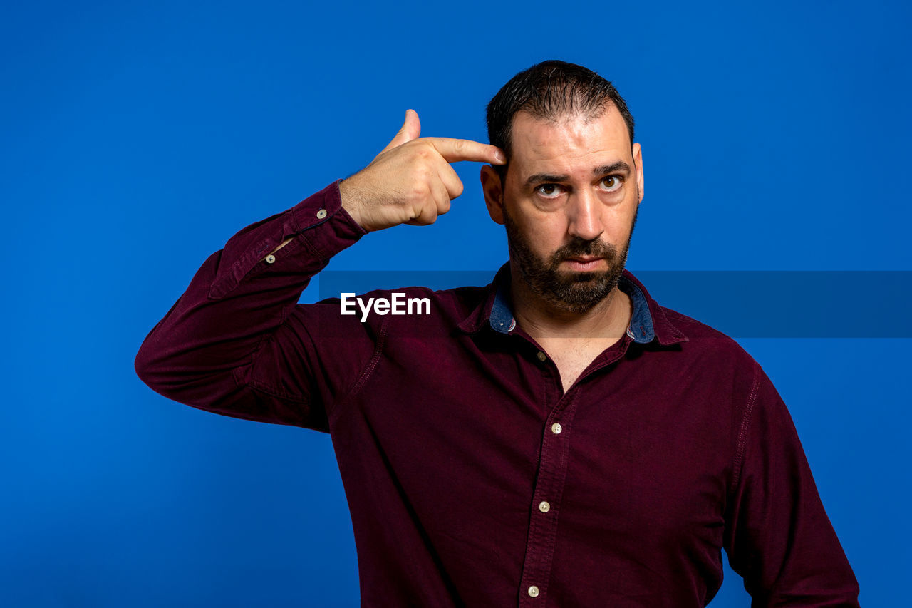 blue, one person, adult, men, blue background, portrait, studio shot, colored background, waist up, beard, looking at camera, facial hair, person, front view, emotion, gesturing, indoors, hand, casual clothing, sign language, looking, polo shirt, young adult, red, standing, clothing, copy space, facial expression