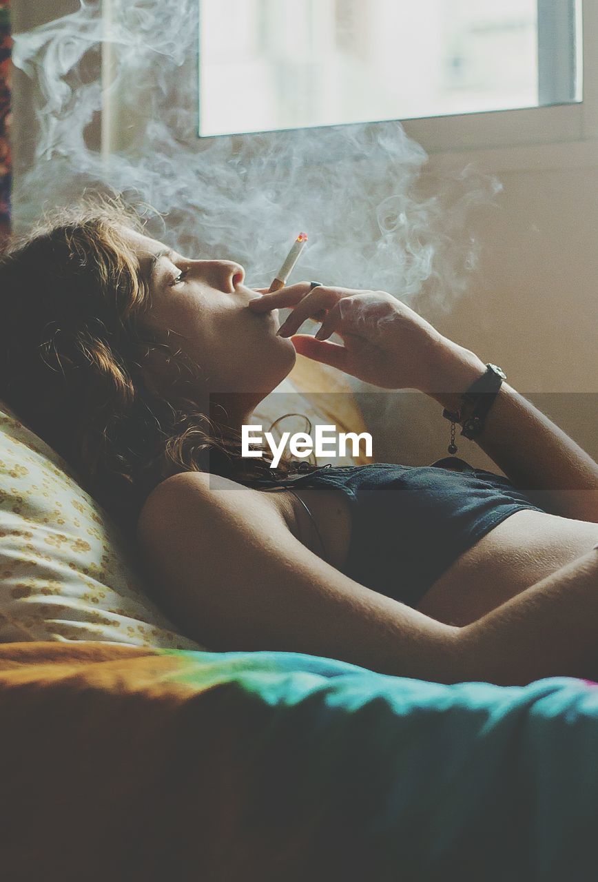 Young woman smoking cigarette while lying on bed