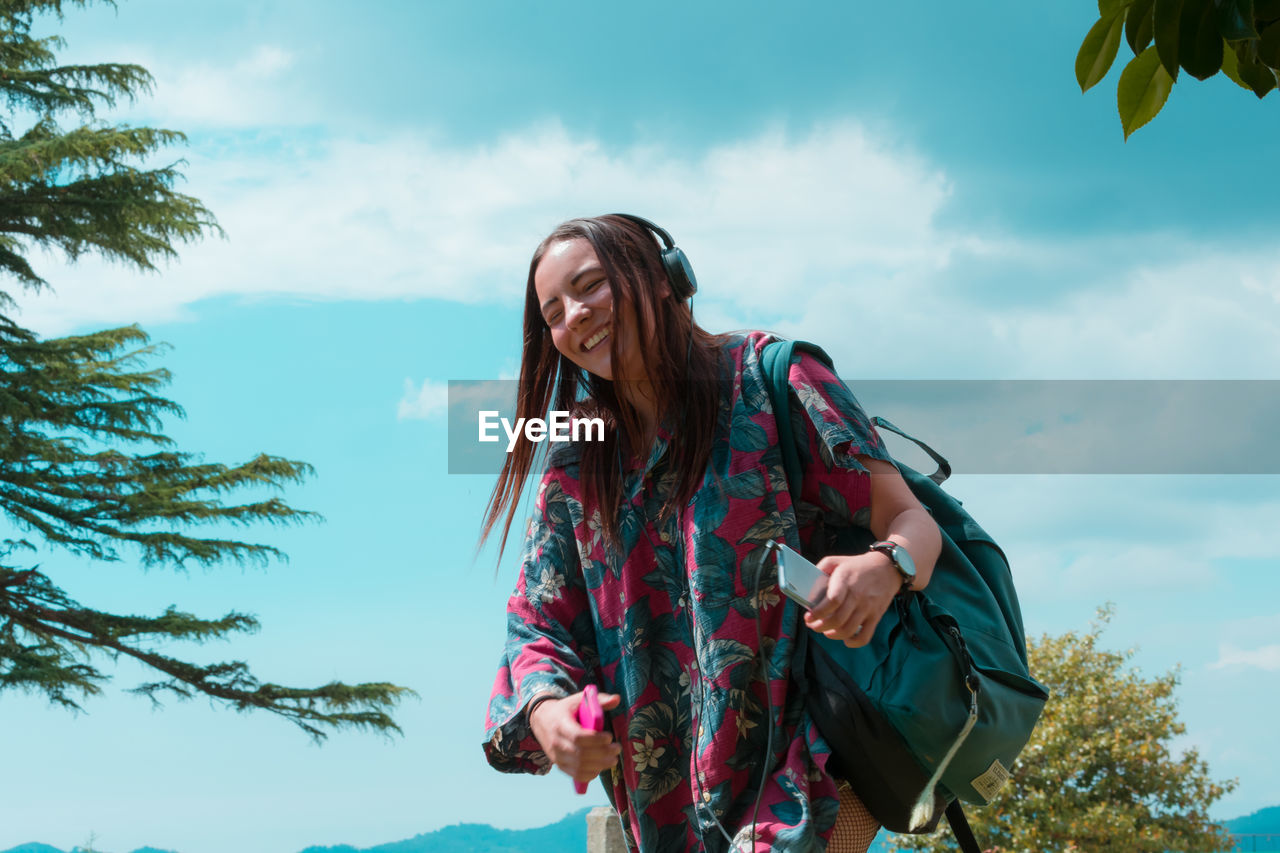 Smiling woman with backpack standing against sky