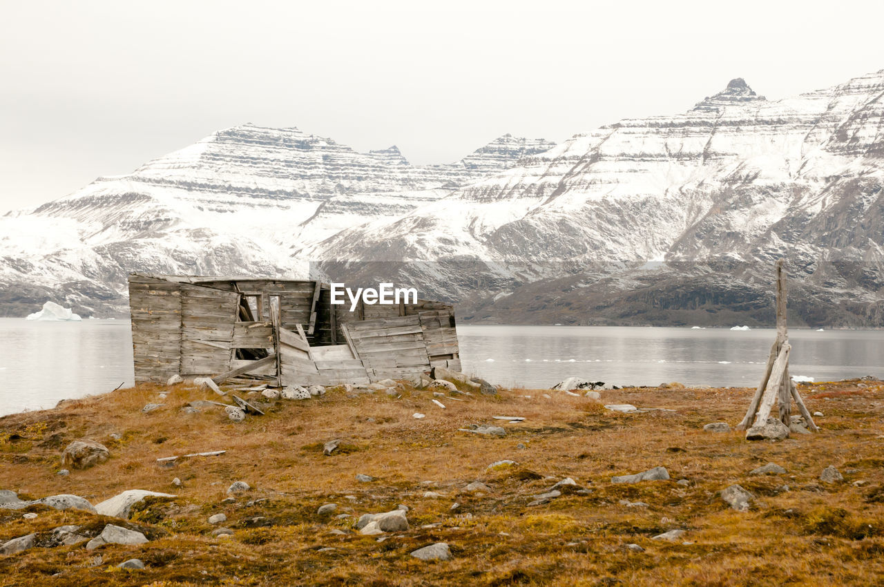 Broken wooden cabin by lake against mountains