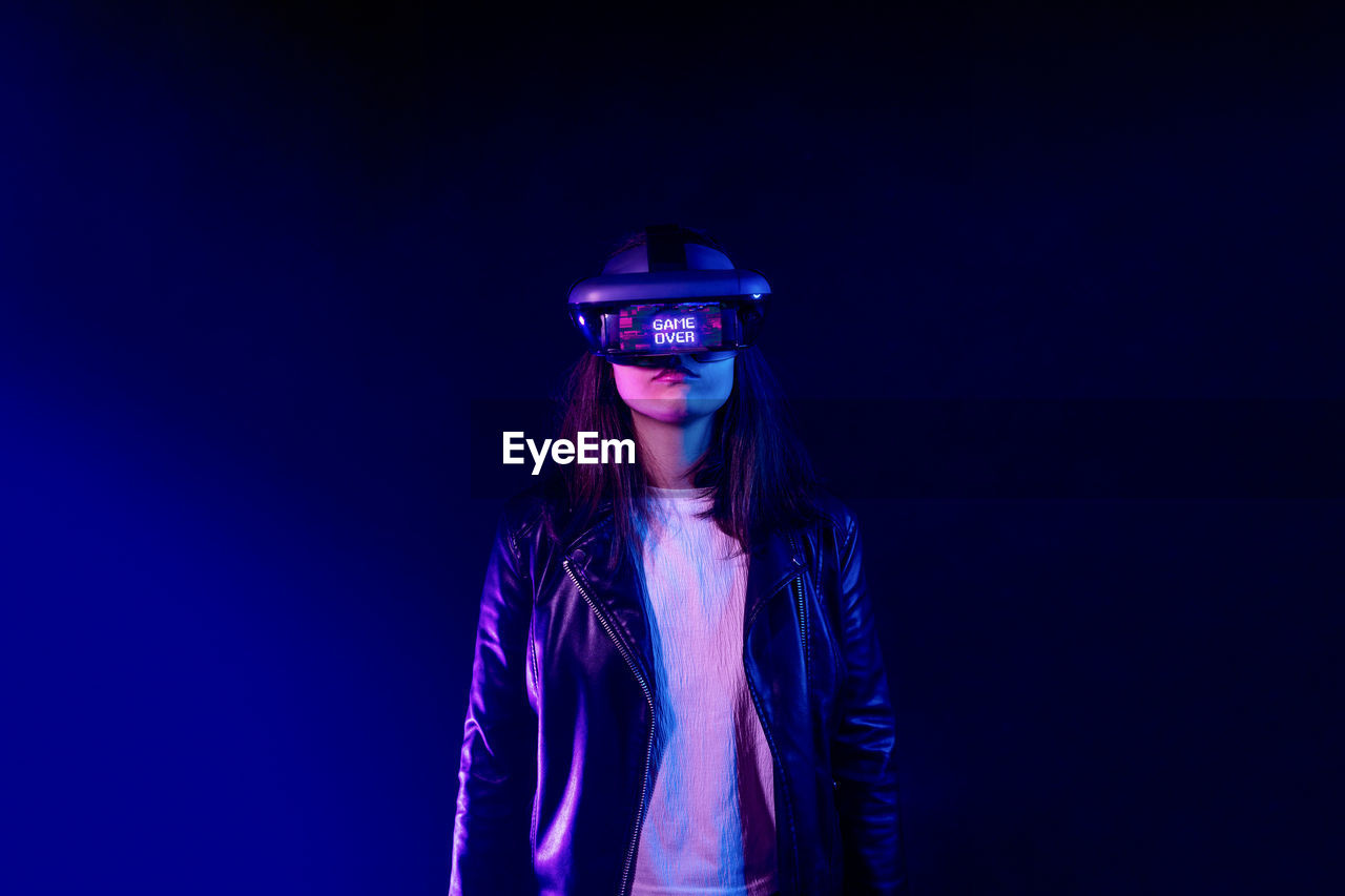 Unrecognizable female wearing modern headset with game over inscription while exploring virtual reality in dark room with neon light near wall
