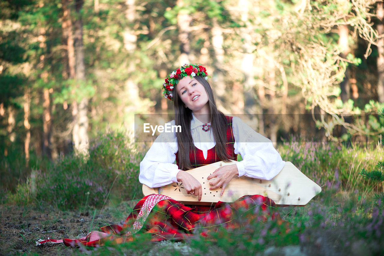 Beautiful musician wearing flowers wreath while playing string instrument at forest