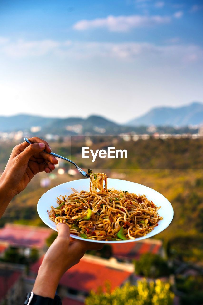 Cropped hand of person holding noodles with chopsticks against mountains