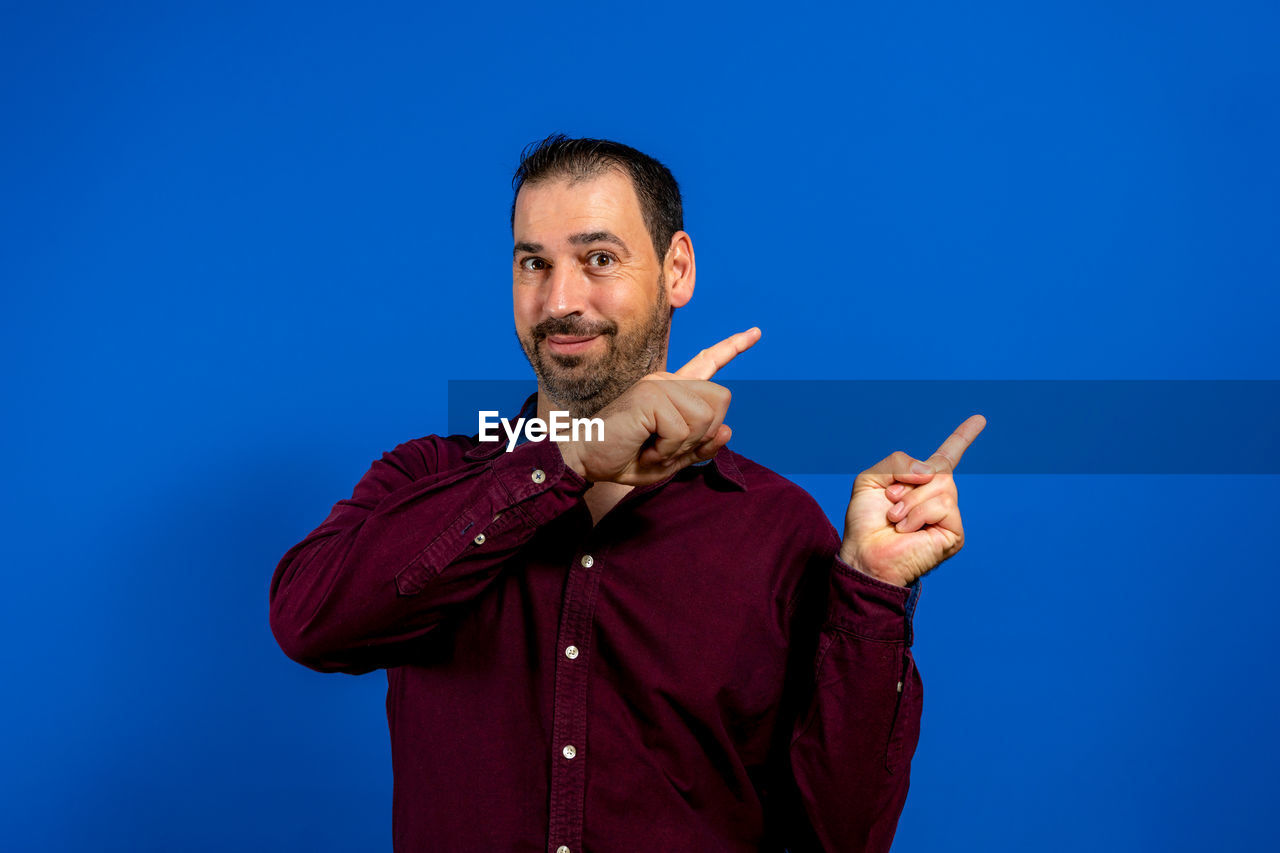 blue, one person, men, adult, gesturing, blue background, colored background, studio shot, sign language, portrait, person, waist up, emotion, looking at camera, smiling, copy space, happiness, hand, beard, facial hair, communication, pointing, indoors, standing, front view, positive emotion, singing, finger, human face, thumbs up, sky, cheerful, hand sign