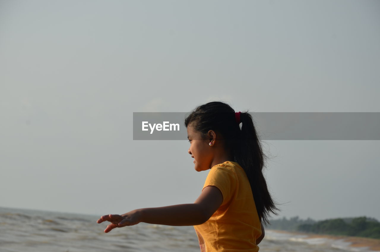 SIDE VIEW OF GIRL STANDING AT BEACH AGAINST SKY