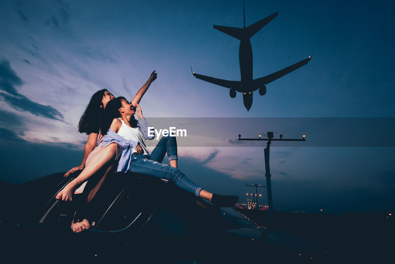 Low angle view of women pointing at airplane landing while sitting on car against sky