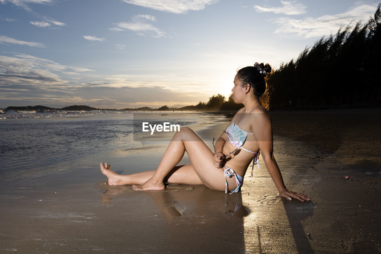 side view of young woman sitting on beach against sky during sunset