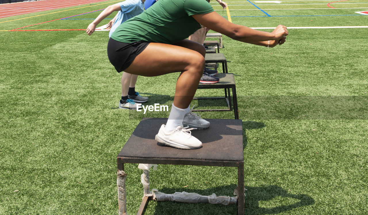 Side view of athletes jumping on different size plyo boxes outdoors on a green turf field.