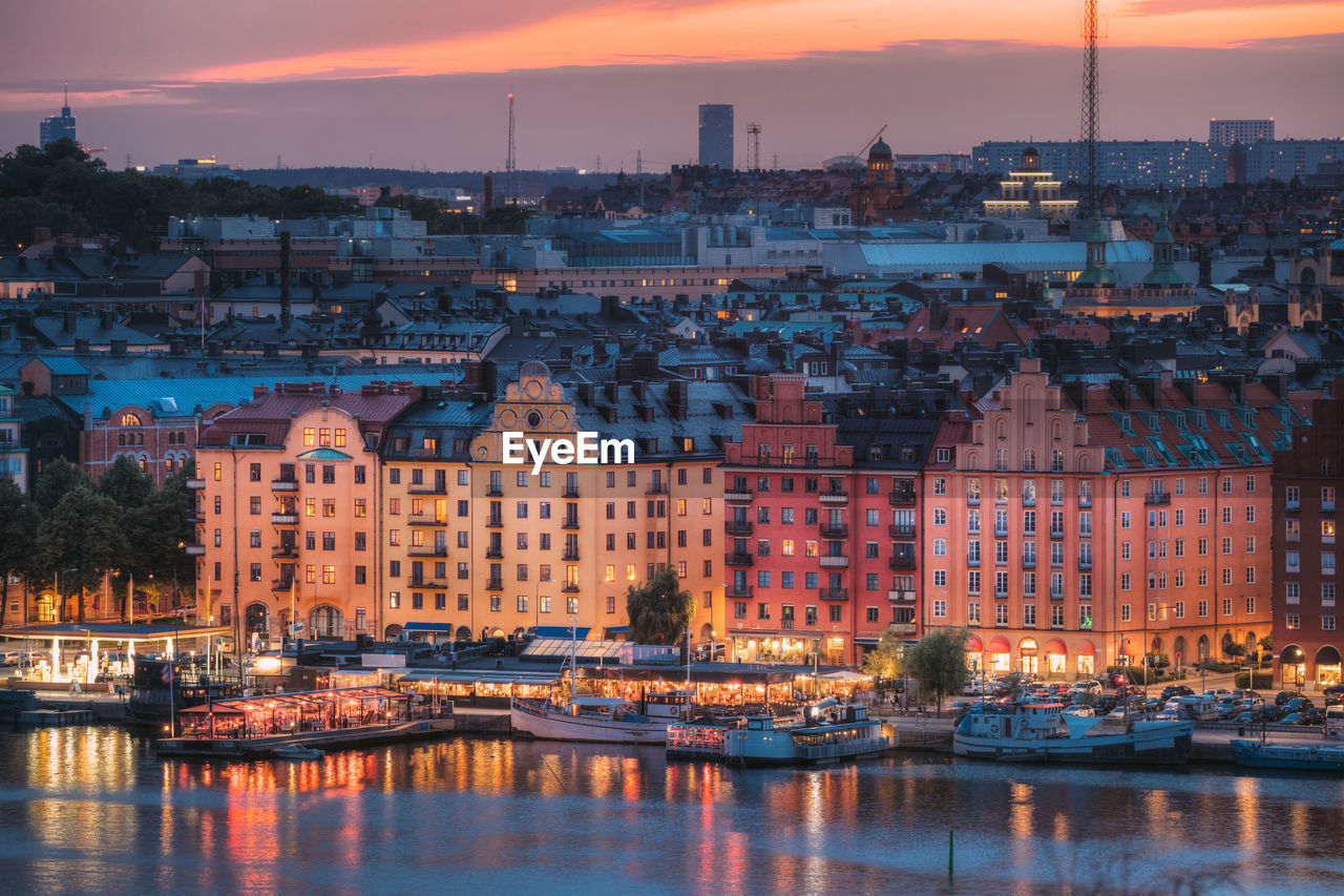 Illuminated buildings by river against sky at sunset