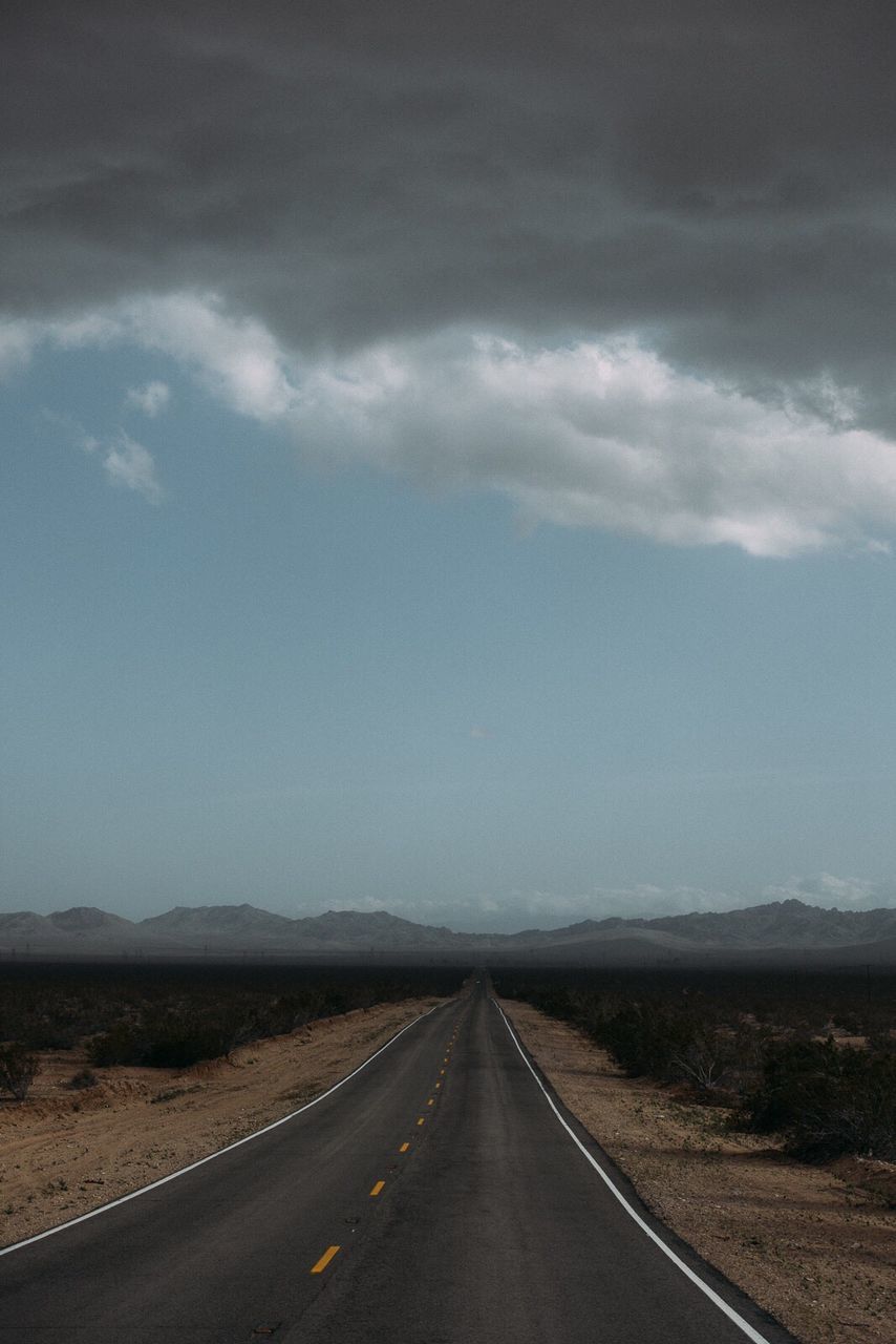 Empty road amidst field by mountains against cloudy sky