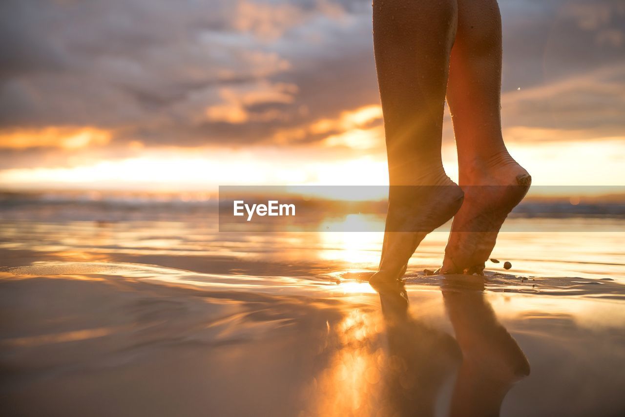 Low section of woman tiptoe standing on shore at beach during sunset