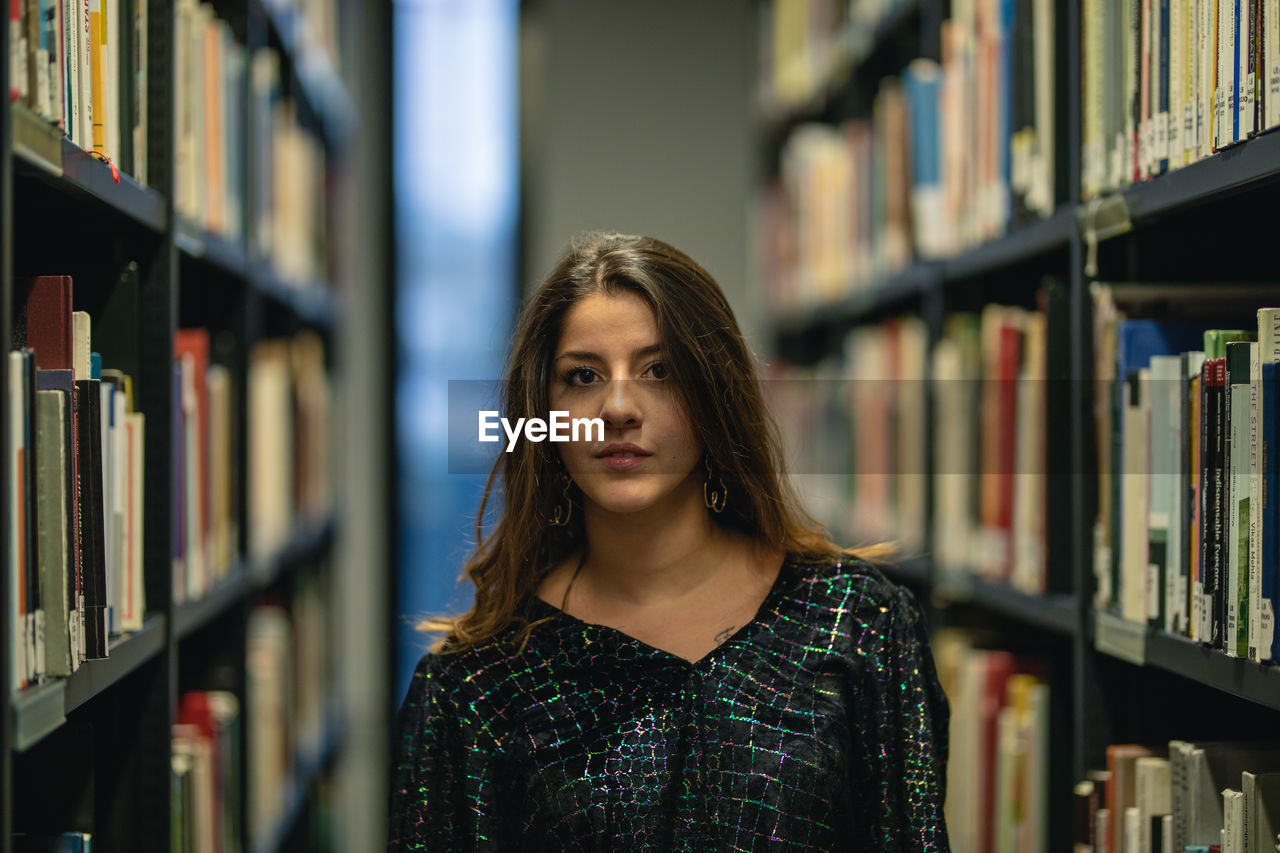 Portrait of young woman standing amidst bookshelf at library