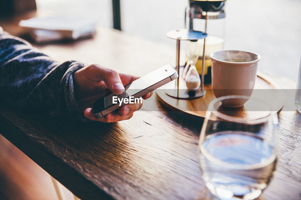 Cropped image of person using mobile phone by tea cup on table