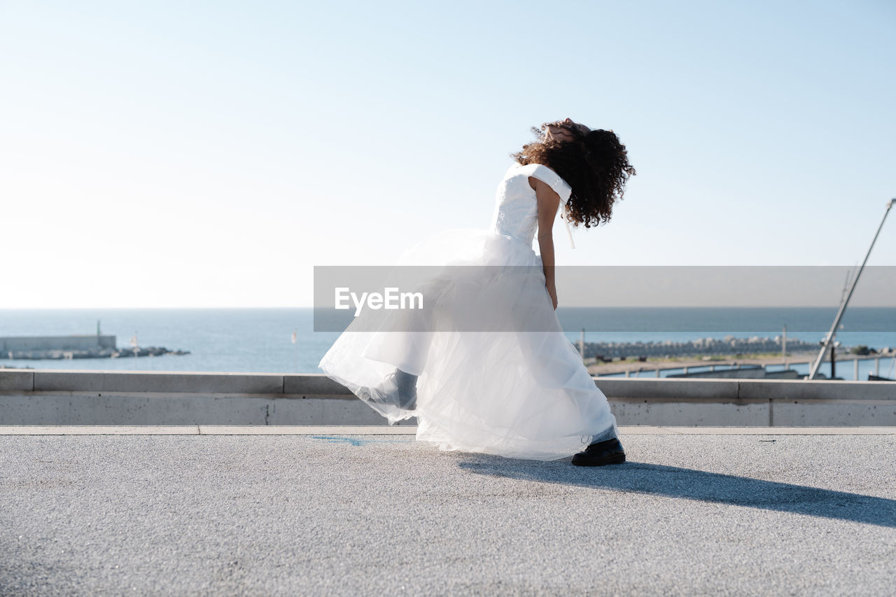 Side view of unrecognizable rebellious female in elegant white dress and brutal boots dance on rooftop in sunlight