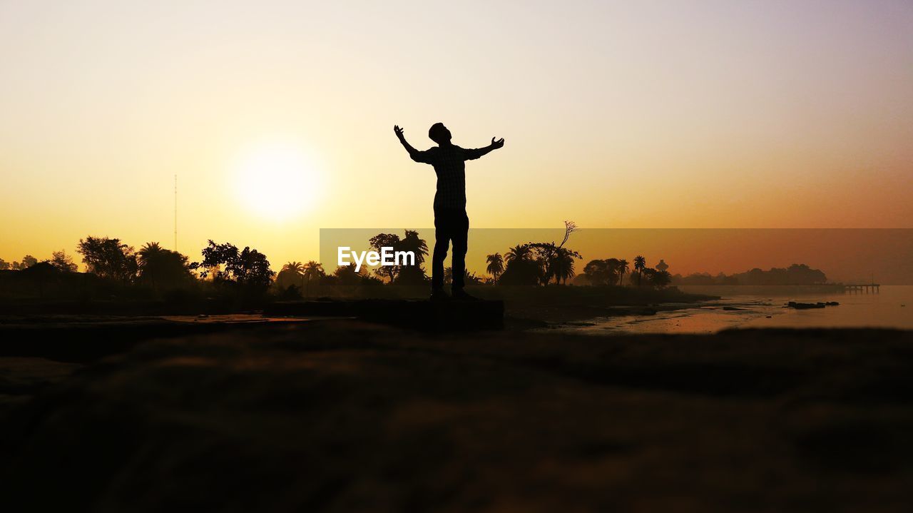 Silhouette man with arms outstretched standing on land against sky during sunset