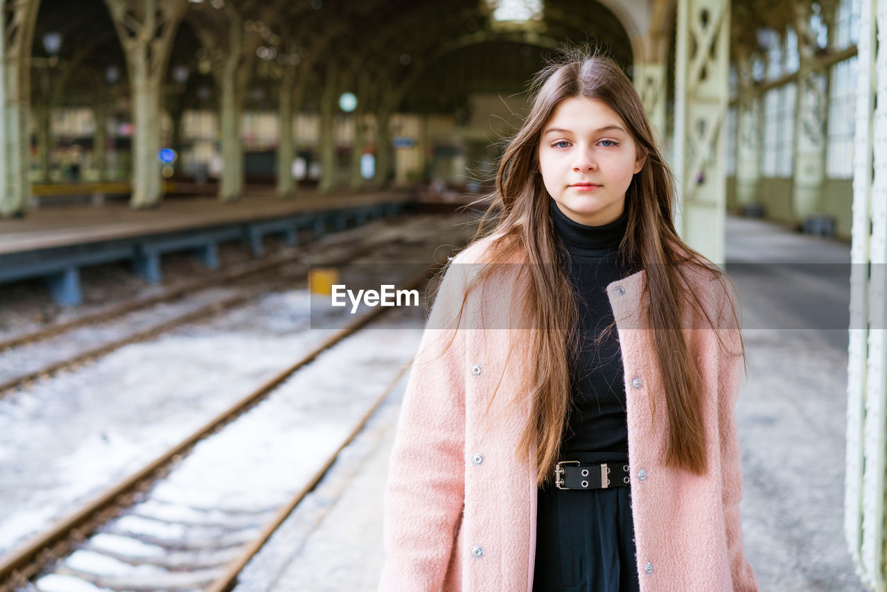 one person, long hair, women, fashion, young adult, hairstyle, portrait, spring, railroad track, track, clothing, adult, standing, architecture, rail transportation, looking at camera, dress, photo shoot, lifestyles, focus on foreground, winter, front view, brown hair, female, nature, city, outdoors, teenager, transportation, day, leisure activity, railroad station, outerwear, casual clothing, emotion, travel, child, waist up, city life, railroad station platform, person, cold temperature
