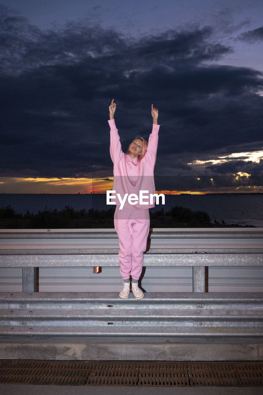 Young woman with pink hair wearing pink track suit shirt standing on road barrier at dusk
