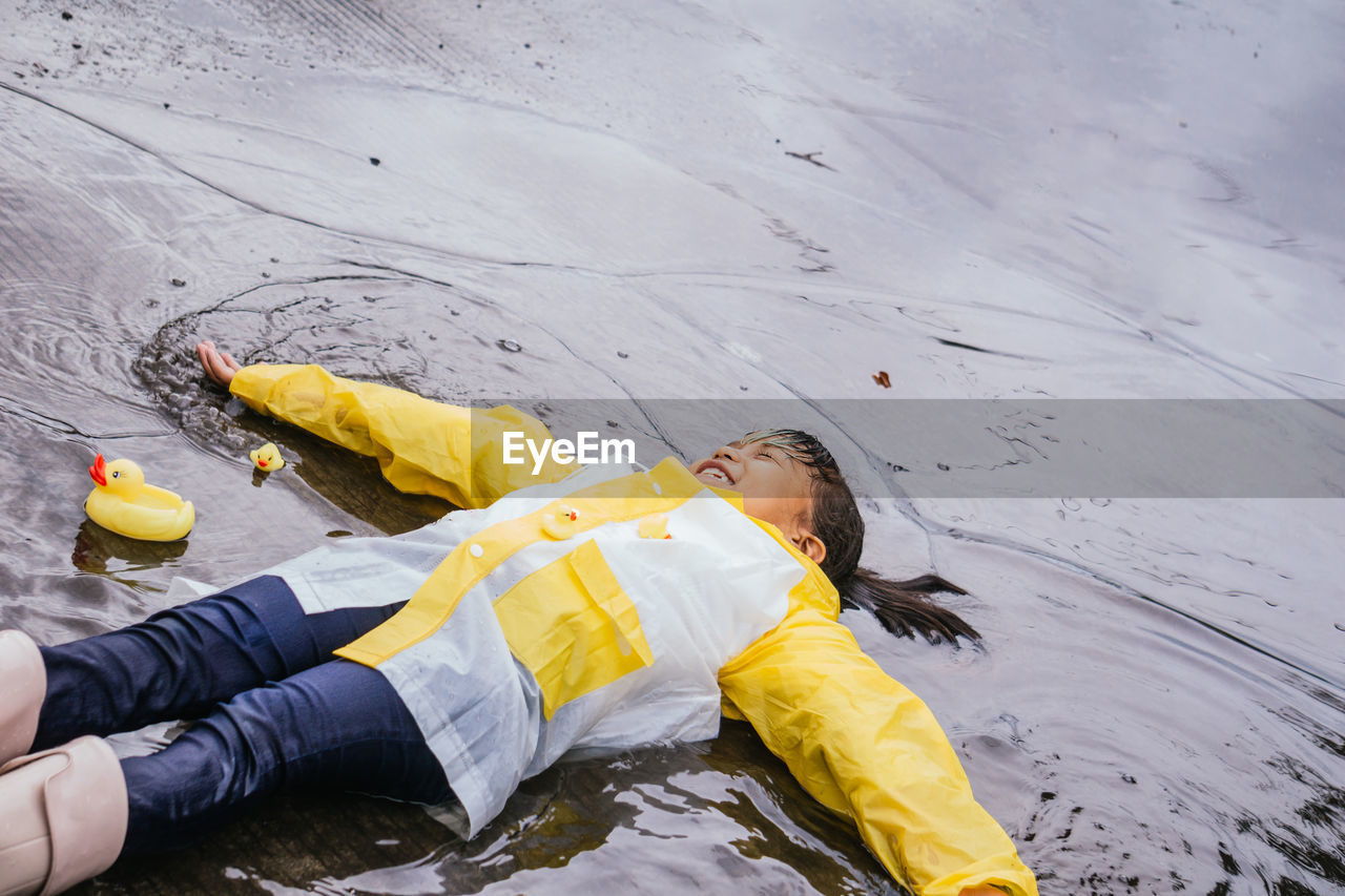 High angle of content asian kid in rubber boots and slicker lying in rippled puddle on rainy day