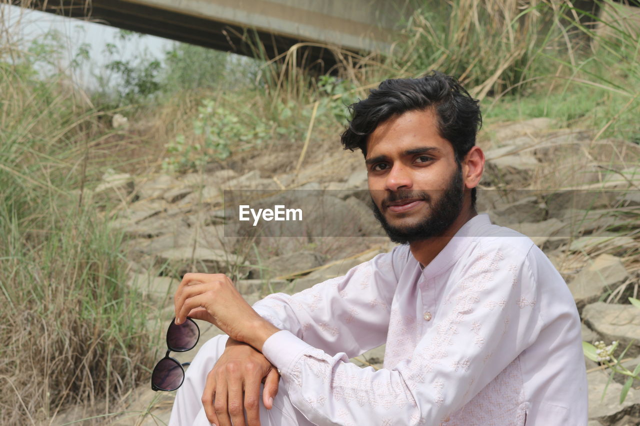 one person, adult, men, young adult, beard, portrait, facial hair, nature, plant, person, looking at camera, sitting, land, smiling, casual clothing, lifestyles, occupation, leisure activity, front view, day, outdoors, button down shirt, happiness, clothing, emotion, landscape, black hair, relaxation, travel, rural scene, grass, holding
