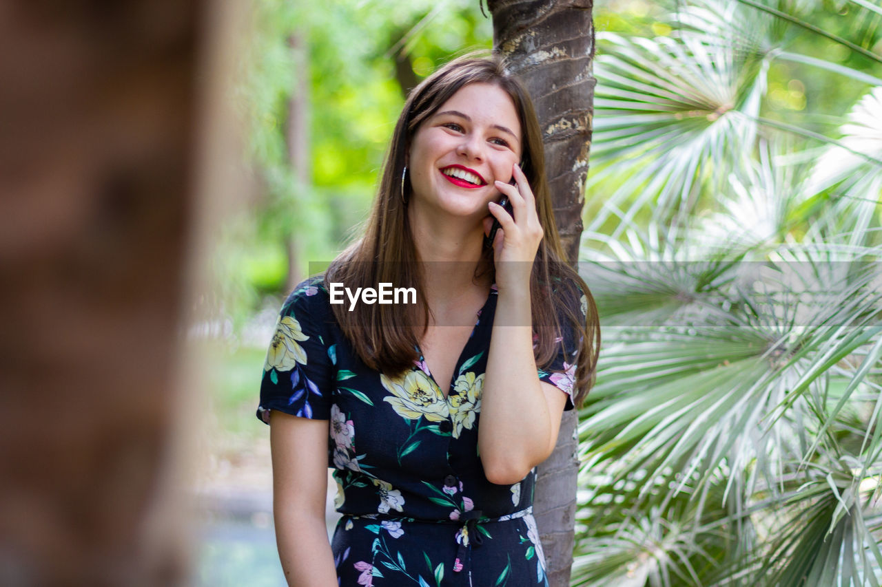 Beautiful young woman using phone while standing against tree trunk