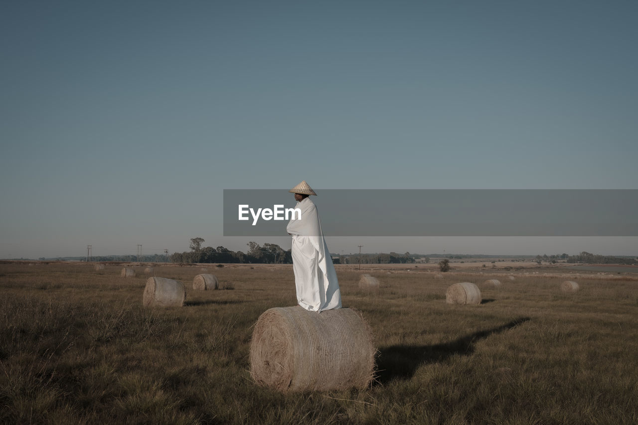 Young man covered with fabric standing on hay bale at agricultural landscape