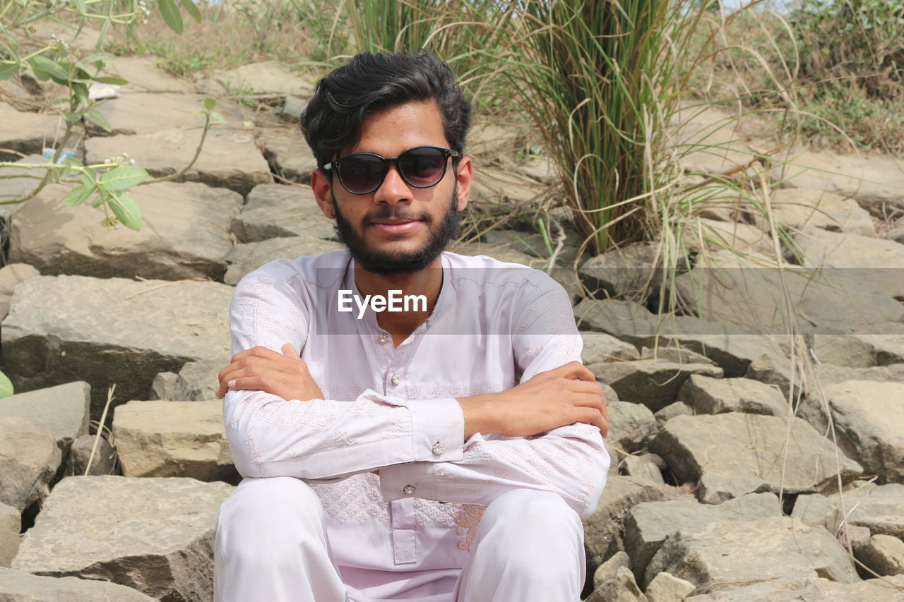 sunglasses, glasses, one person, fashion, sitting, front view, young adult, leisure activity, adult, rock, casual clothing, men, nature, portrait, beard, lifestyles, facial hair, day, relaxation, looking at camera, person, land, sunlight, three quarter length, clothing, outdoors, smiling, vacation, trip, human face, black hair