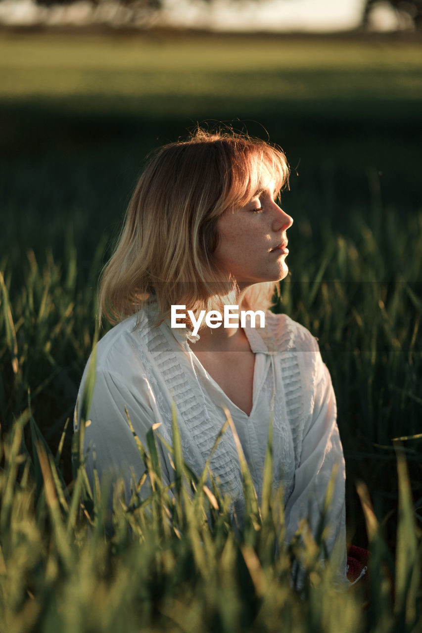 Peaceful young female in retro styled white blouse sitting amidst tall green grass and closed eyes while resting in summer evening in countryside