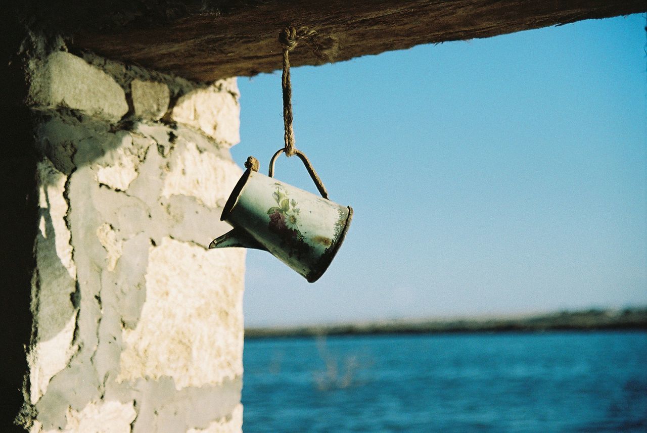 Watering can hanging on rope at abandoned building by sea against blue sky