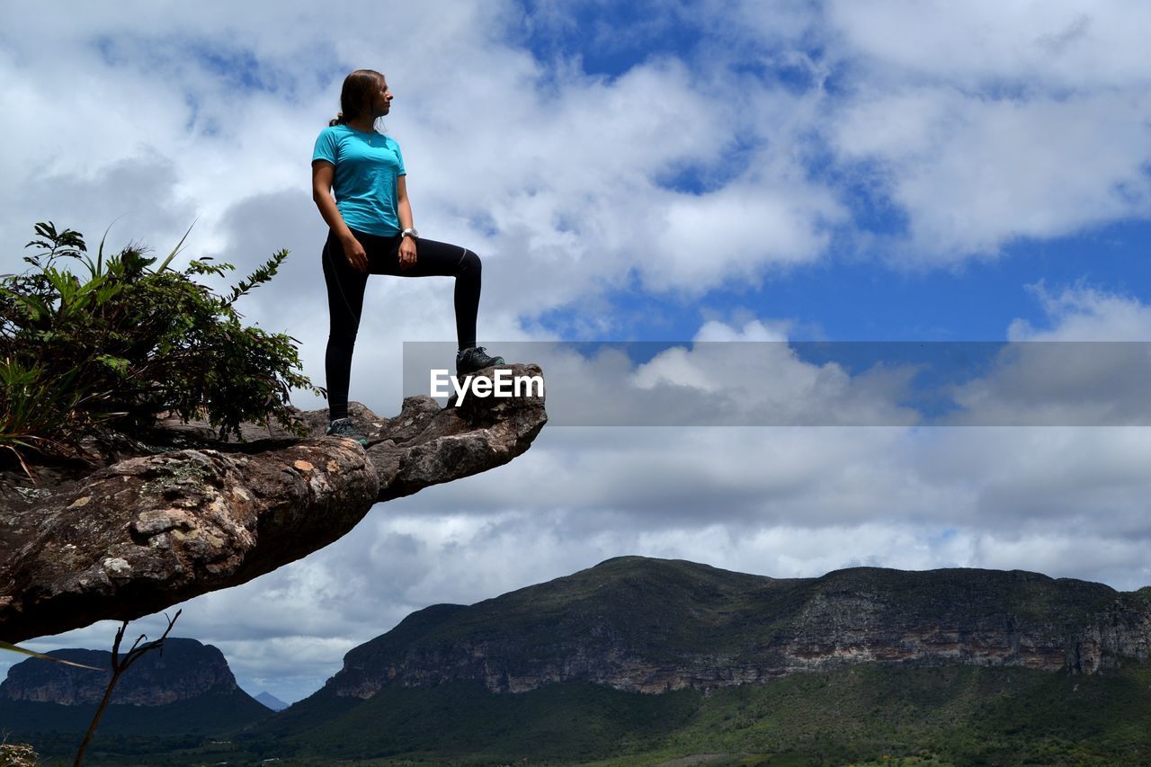 Low angle view of young woman standing on cliff against cloudy sky