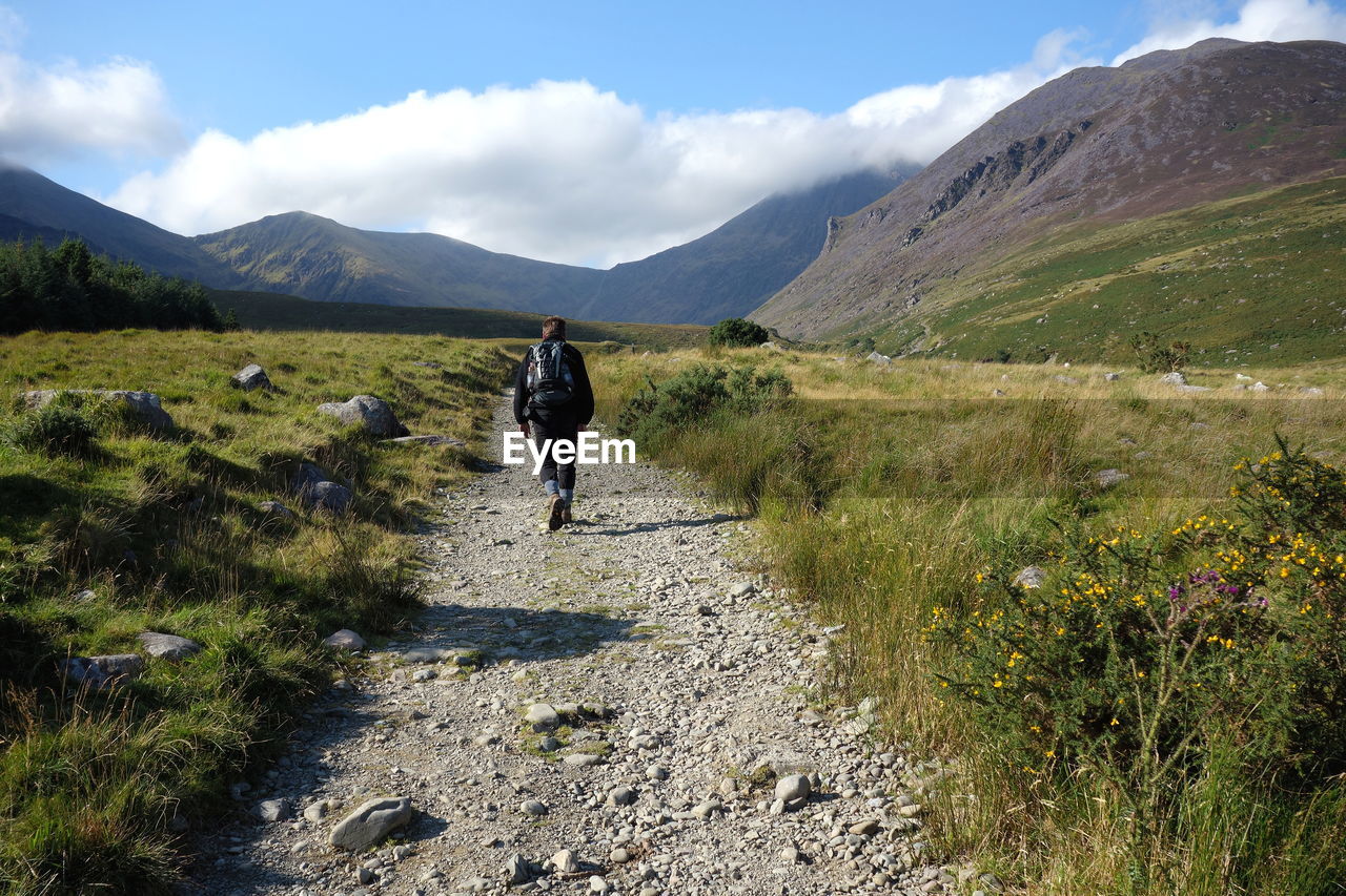 Rear view of man walking on trail leading towards mountains against sky