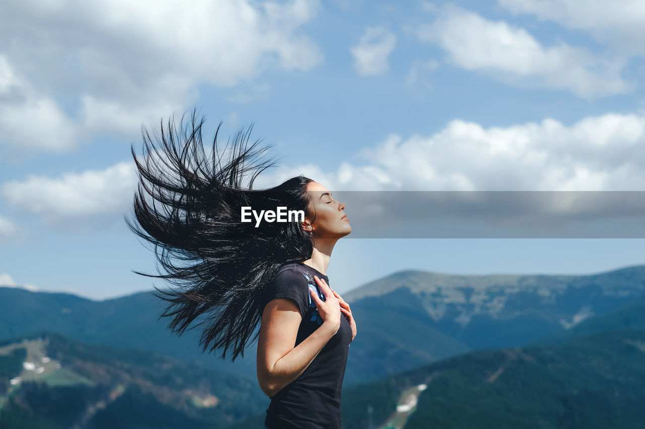 Side view of mid adult woman with eyes closed standing on mountain against cloudy sky