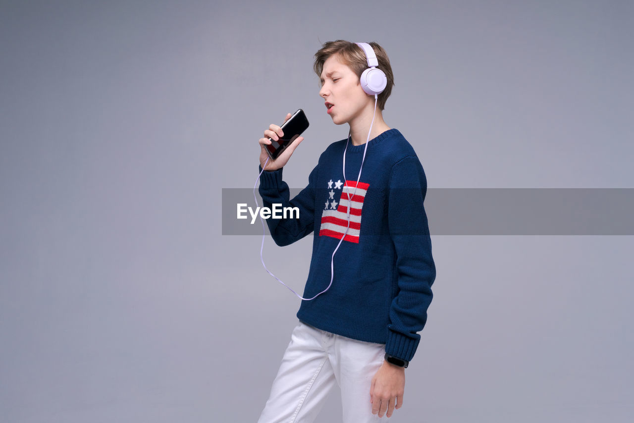 one person, studio shot, headphones, adult, young adult, listening, standing, sports, communication, indoors, clothing, music, athlete, technology, holding, person, blue, waist up, copy space, emotion, gray, wireless technology, portrait, gray background, arts culture and entertainment, looking, lifestyles, three quarter length, colored background
