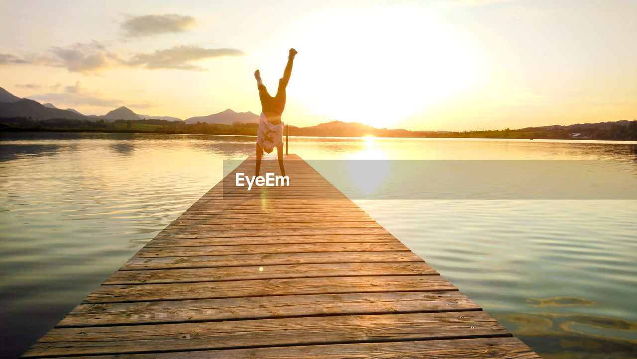 Woman doing handstand on pier over lake during sunset