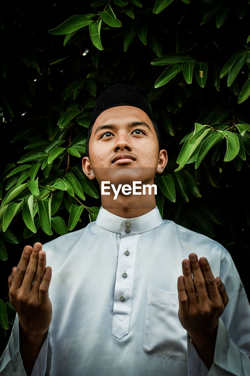 Young man looking up while praying against tree
