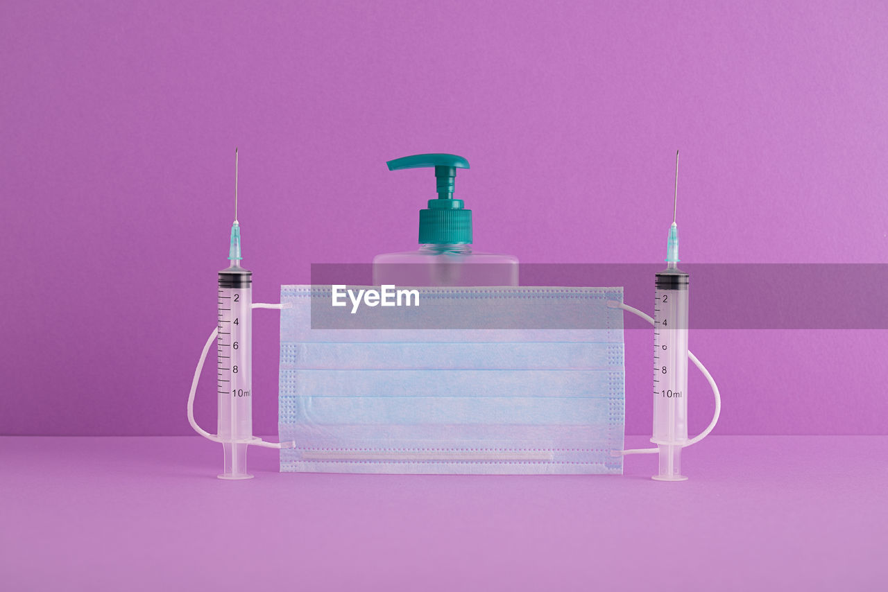 Syringe and face mask with hand sanitizer on purple background