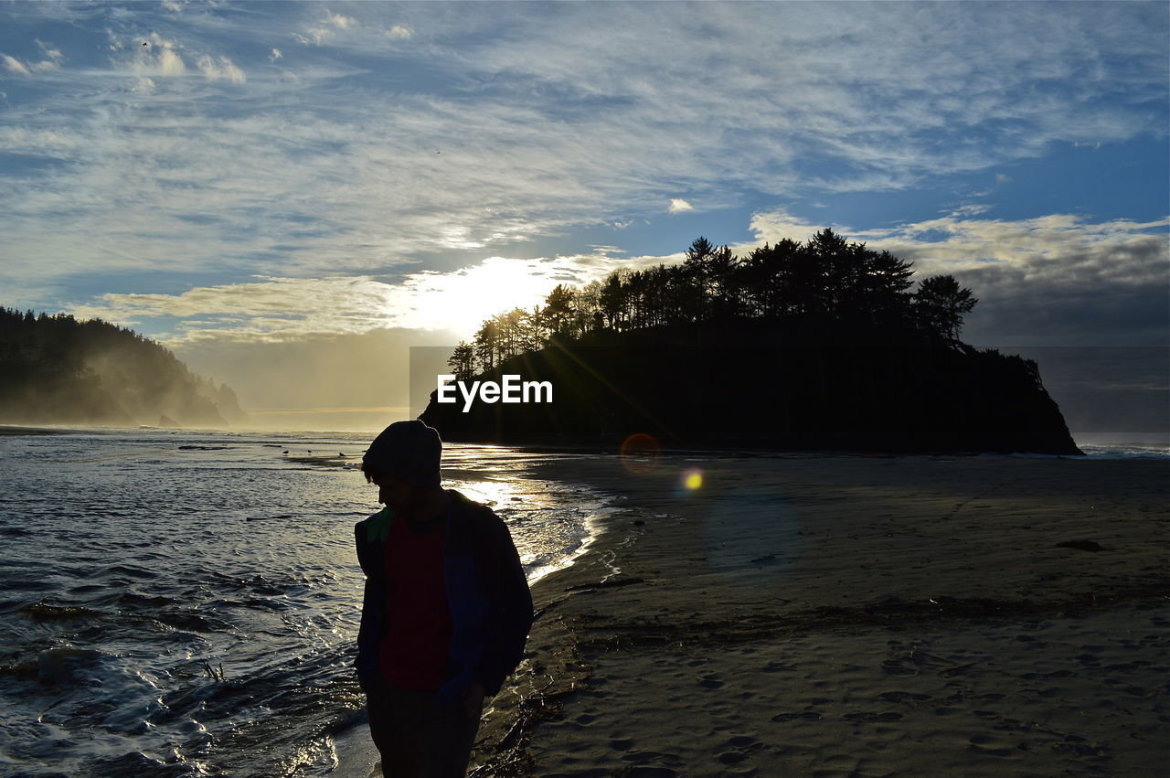 Man walking on shore against cloudy sky during sunset