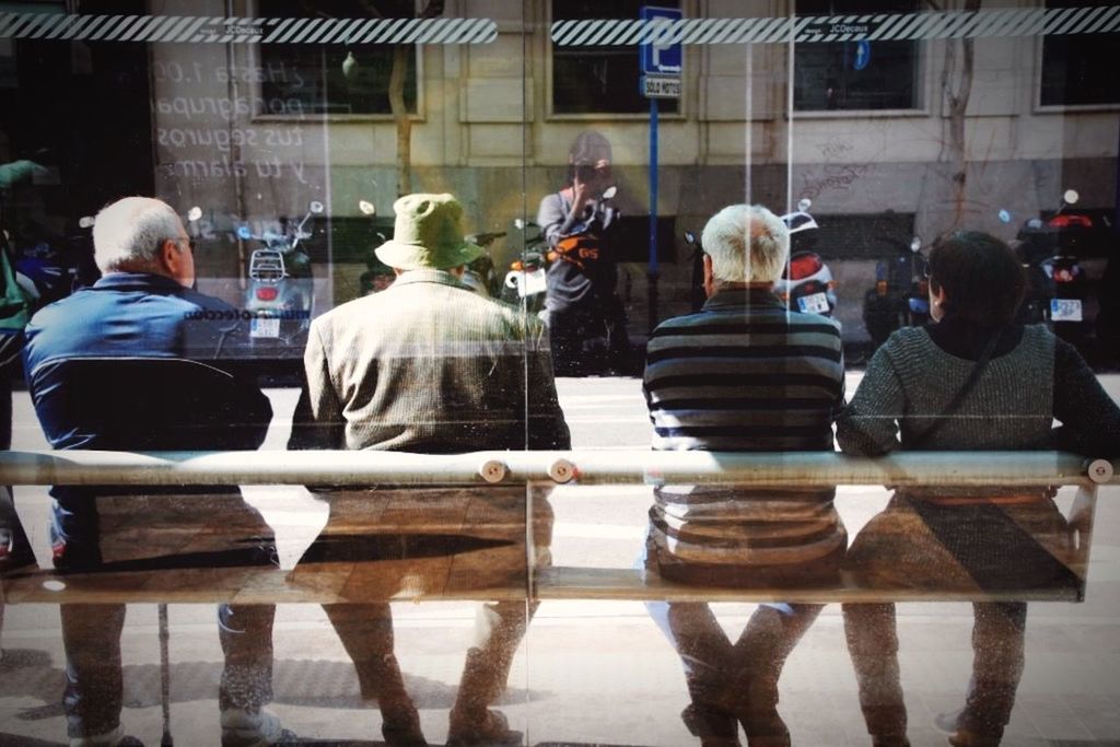 Reflection of woman photographing people sitting on bench on glass