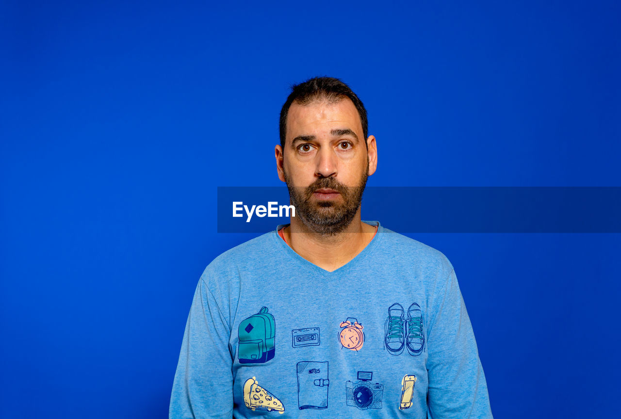 blue, one person, blue background, portrait, studio shot, colored background, front view, adult, beard, facial hair, looking at camera, men, t-shirt, indoors, copy space, person, waist up, clothing, casual clothing, standing, young adult, individuality