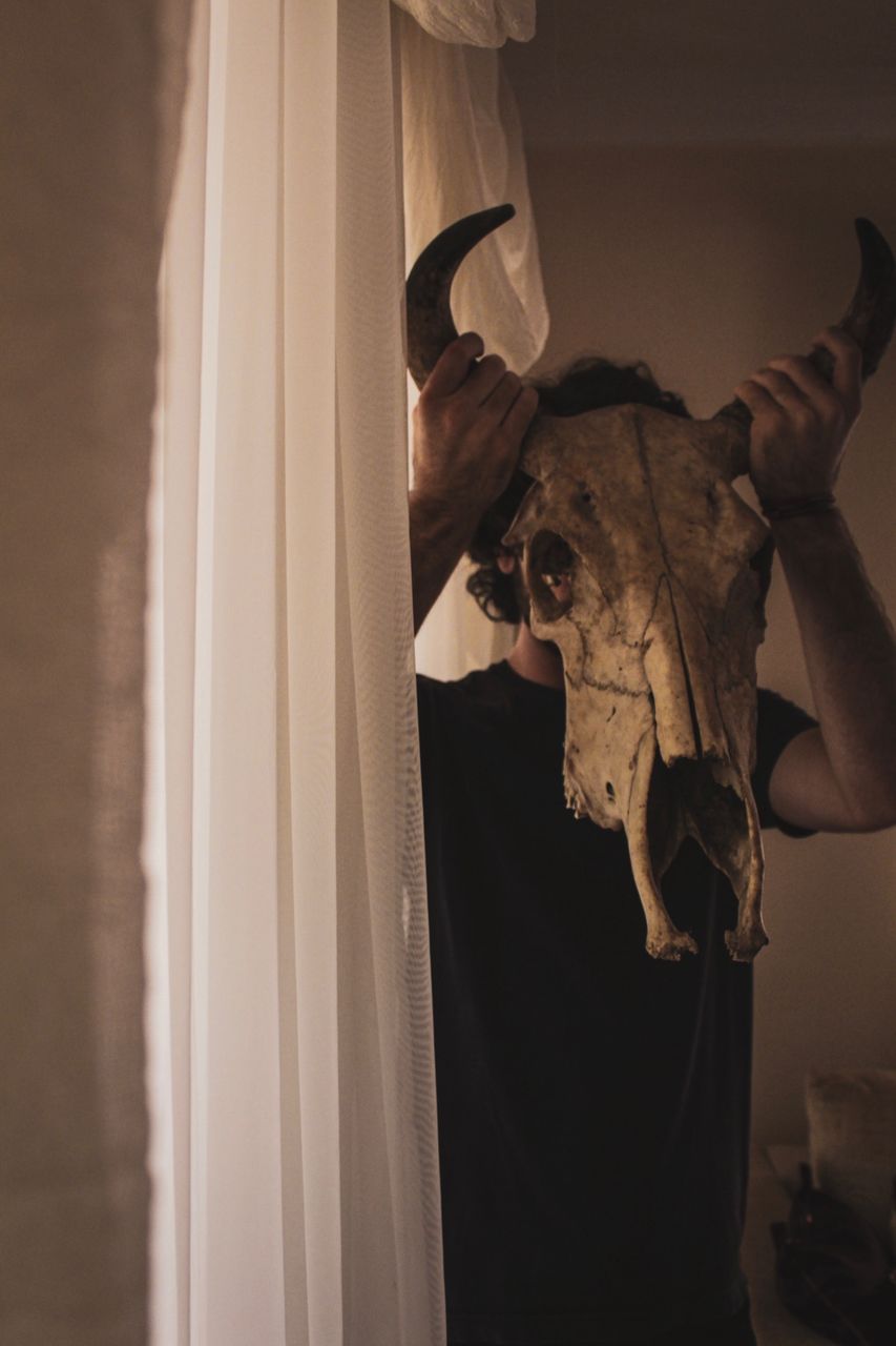 Man holding animal skull over face while standing by window at home