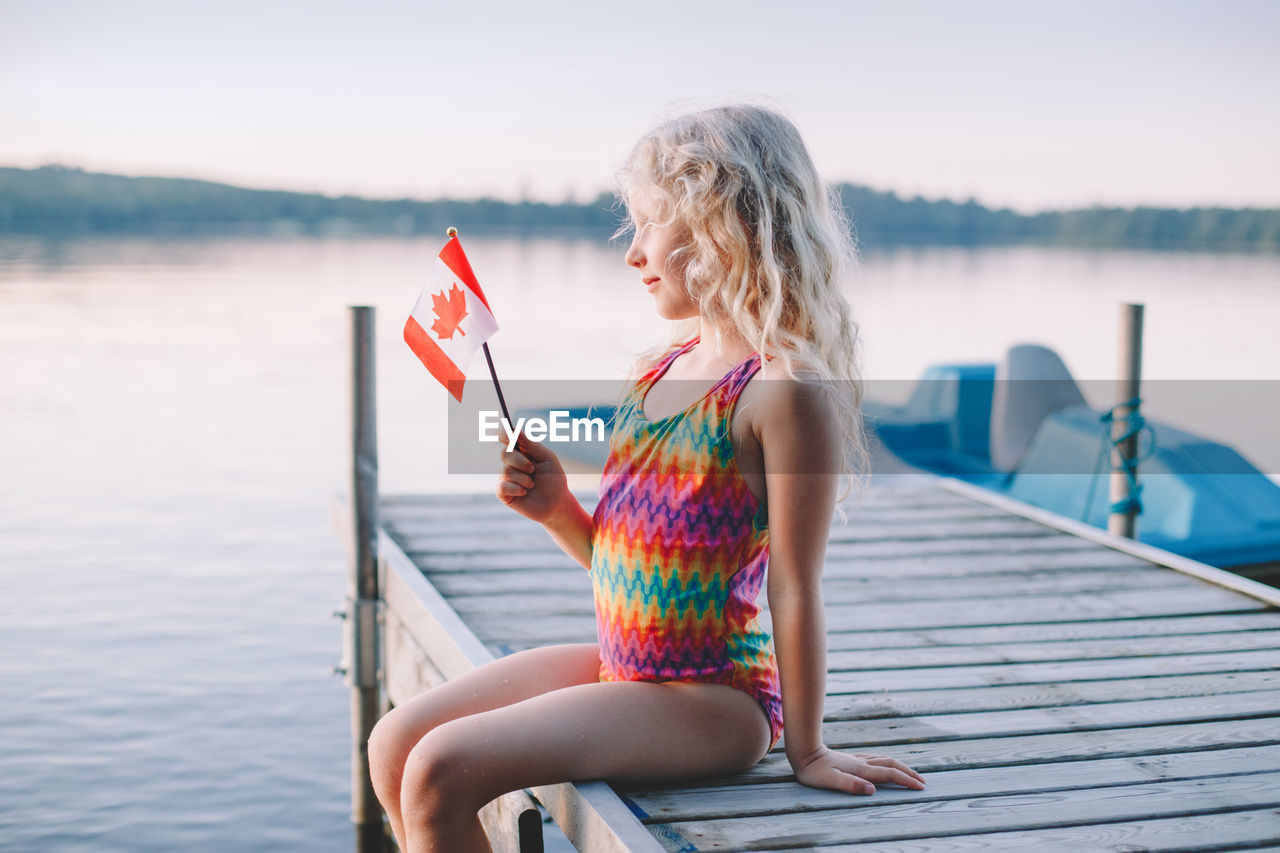 Girl sitting on pier by lake and waving canadian flag. kid celebrating canada day holiday outdoor