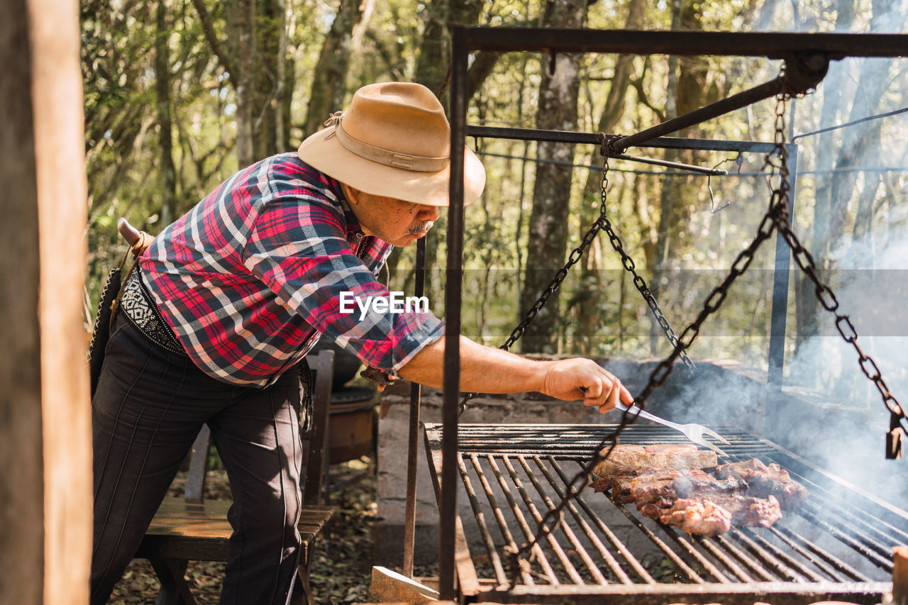 Side view of argentinian male in hat with fork adjusting fresh meat pieces on rack over flame in garden