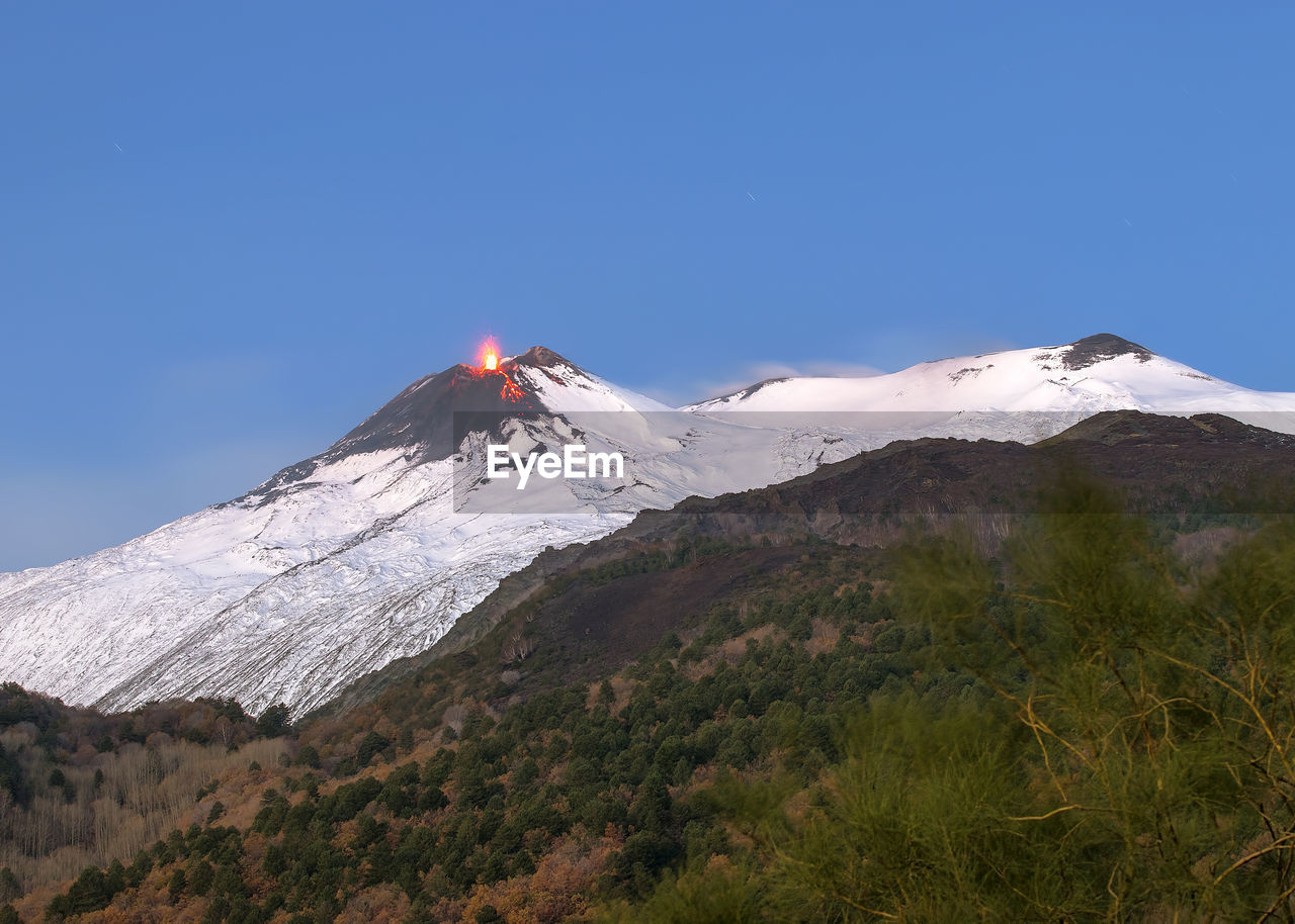 Low angle view of snowcapped volcano erupting against blue sky