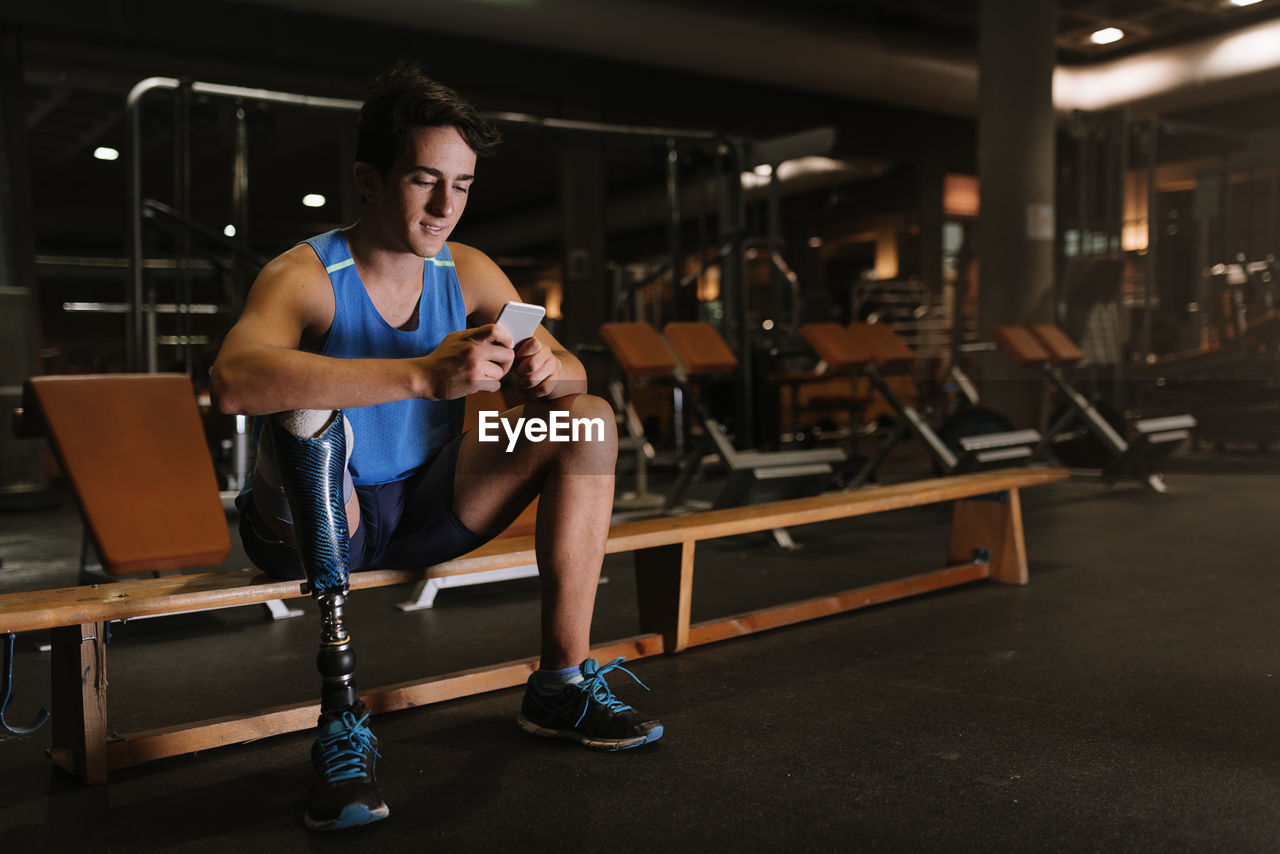 Full length of young man with prosthetic leg using phone while sitting on bench in gym