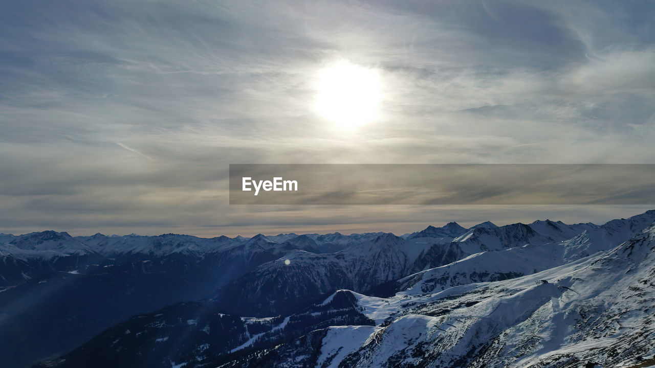 sky, scenics - nature, snow, environment, cold temperature, winter, beauty in nature, mountain, cloud, landscape, sun, nature, mountain range, sunlight, tranquility, tranquil scene, sunbeam, no people, snowcapped mountain, travel destinations, travel, non-urban scene, outdoors, idyllic, dramatic sky, sunset, ice, lens flare, blue, mountain peak, tourism, extreme terrain, frozen, land