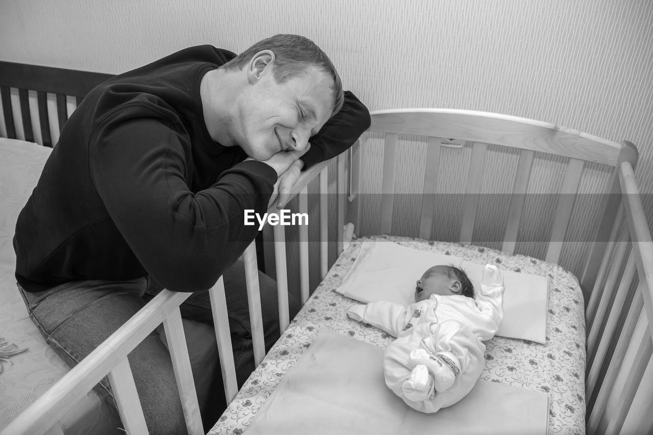 Baby in crib while happy man sitting on bed at home